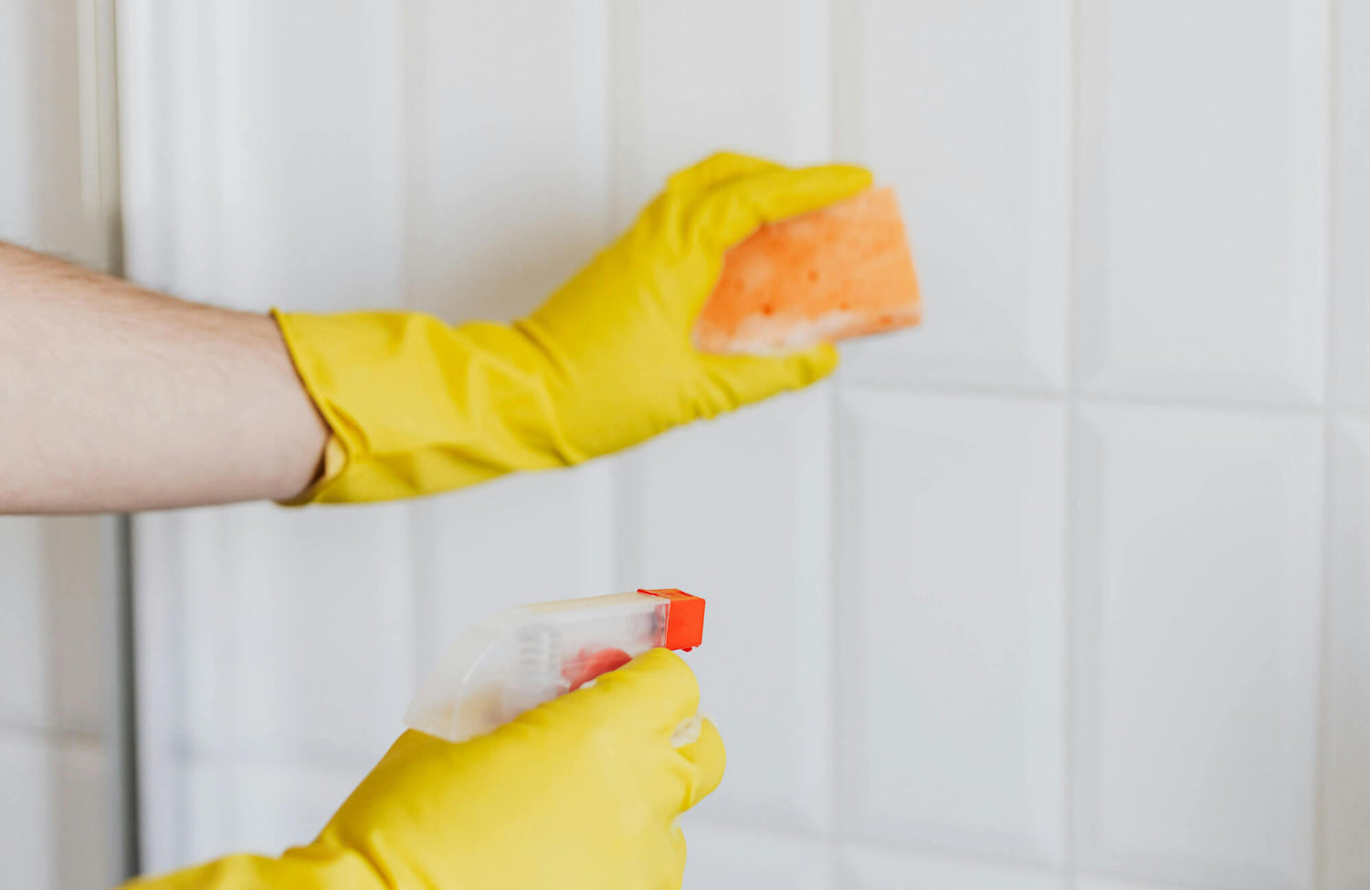 Hands in yellow cleaning gloves scrubbing glossy white subway tiles with an orange sponge and spray cleaner, ensuring a sparkling finish.