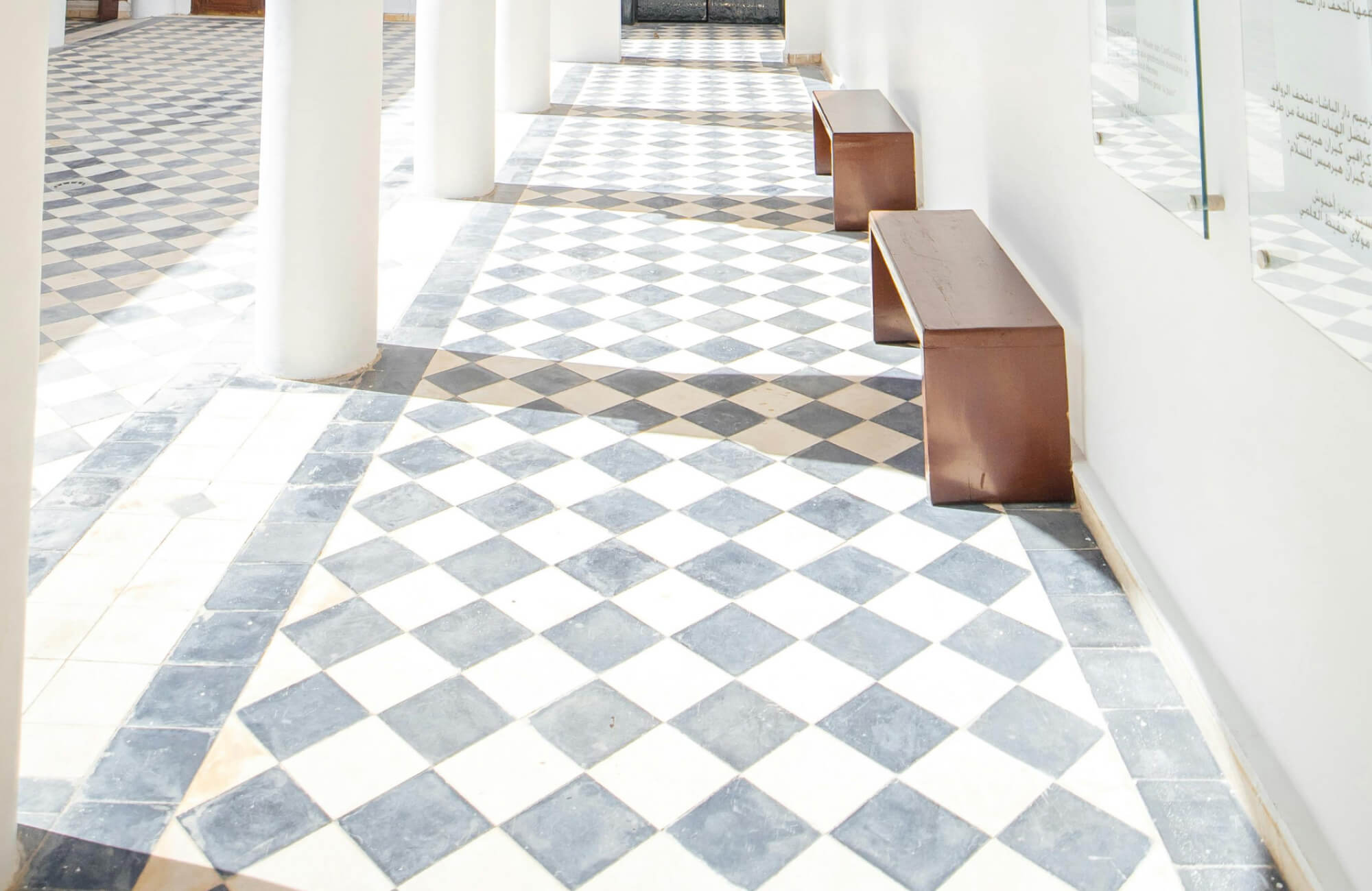 Bright hallway featuring classic black and white checkered floor tiles, with modern wooden benches and white columns enhancing the space’s airy feel.