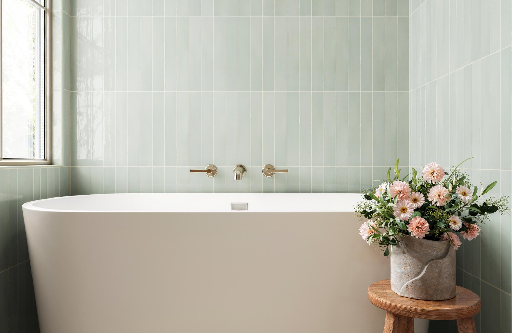 Soft green subway tiles accentuate a minimalist bathroom, with a freestanding white bathtub and fresh floral arrangement adding elegance.