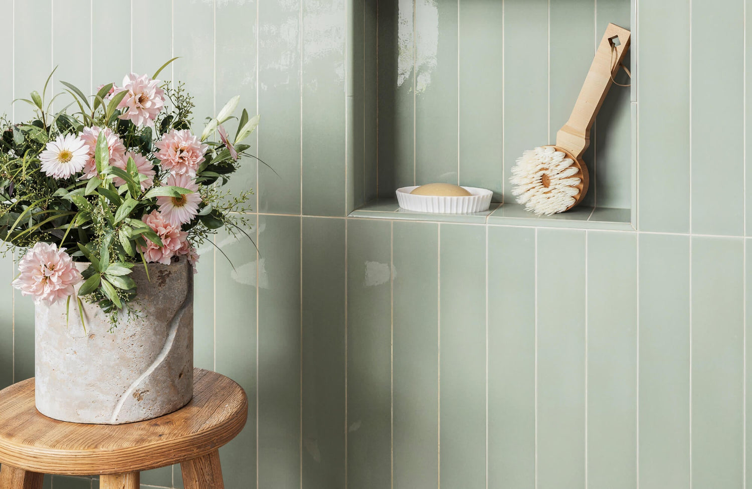 Soft green vertical subway tiles with a built-in shelf holding a wooden bath brush and soap, complemented by a vase of fresh pink flowers on a wooden stool.