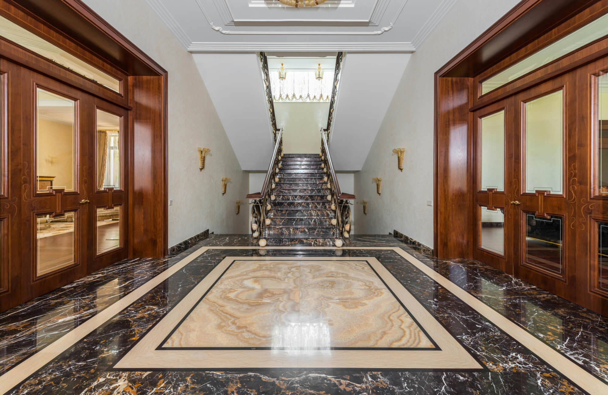 Grand entrance hall featuring luxurious marble flooring with intricate inlays and a sweeping staircase, framed by rich wooden doors.