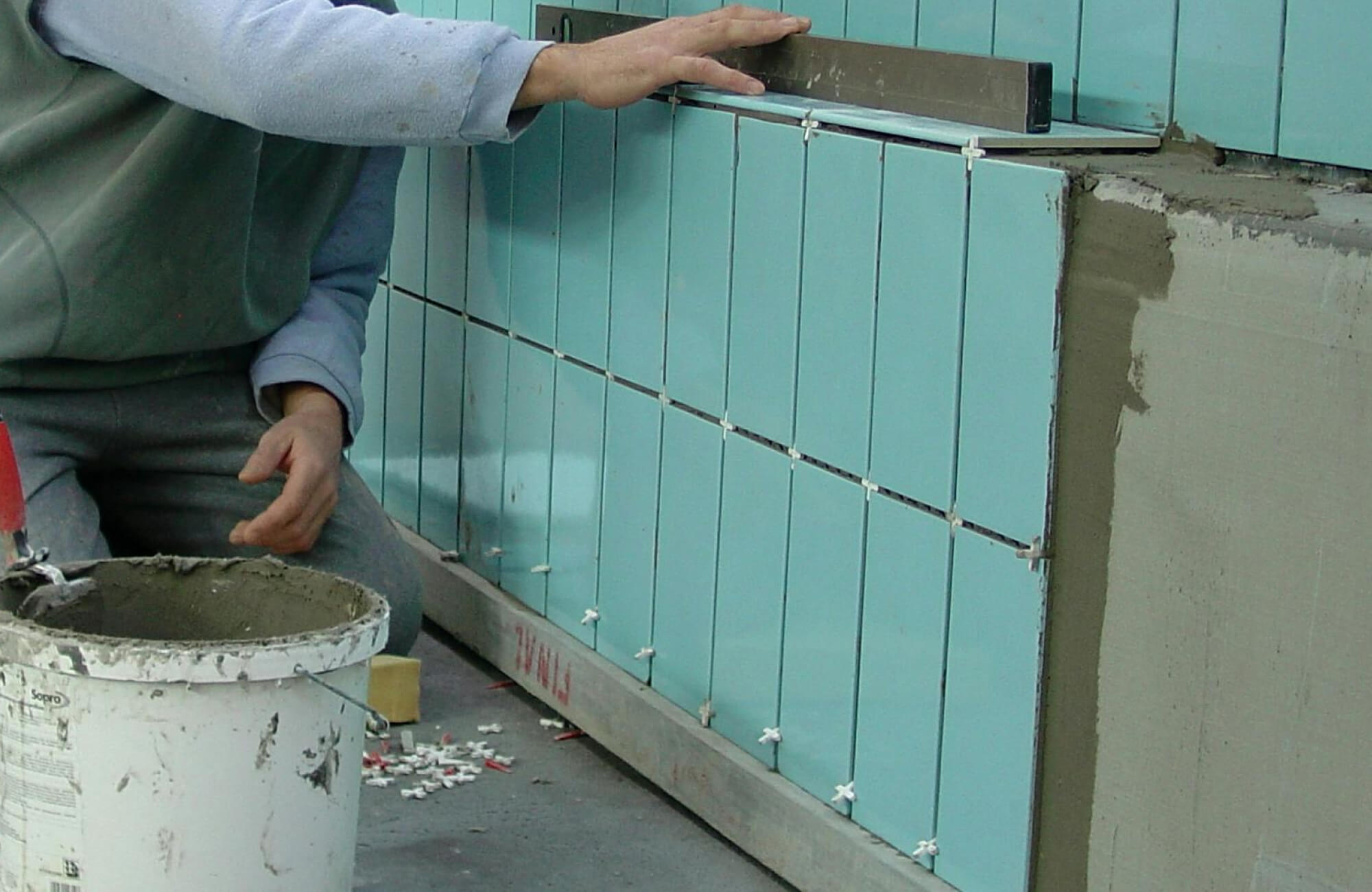 Skilled worker aligning glossy aqua-blue subway tiles on a wall, with precision tools and adhesive bucket nearby, creating a sleek modern finish.