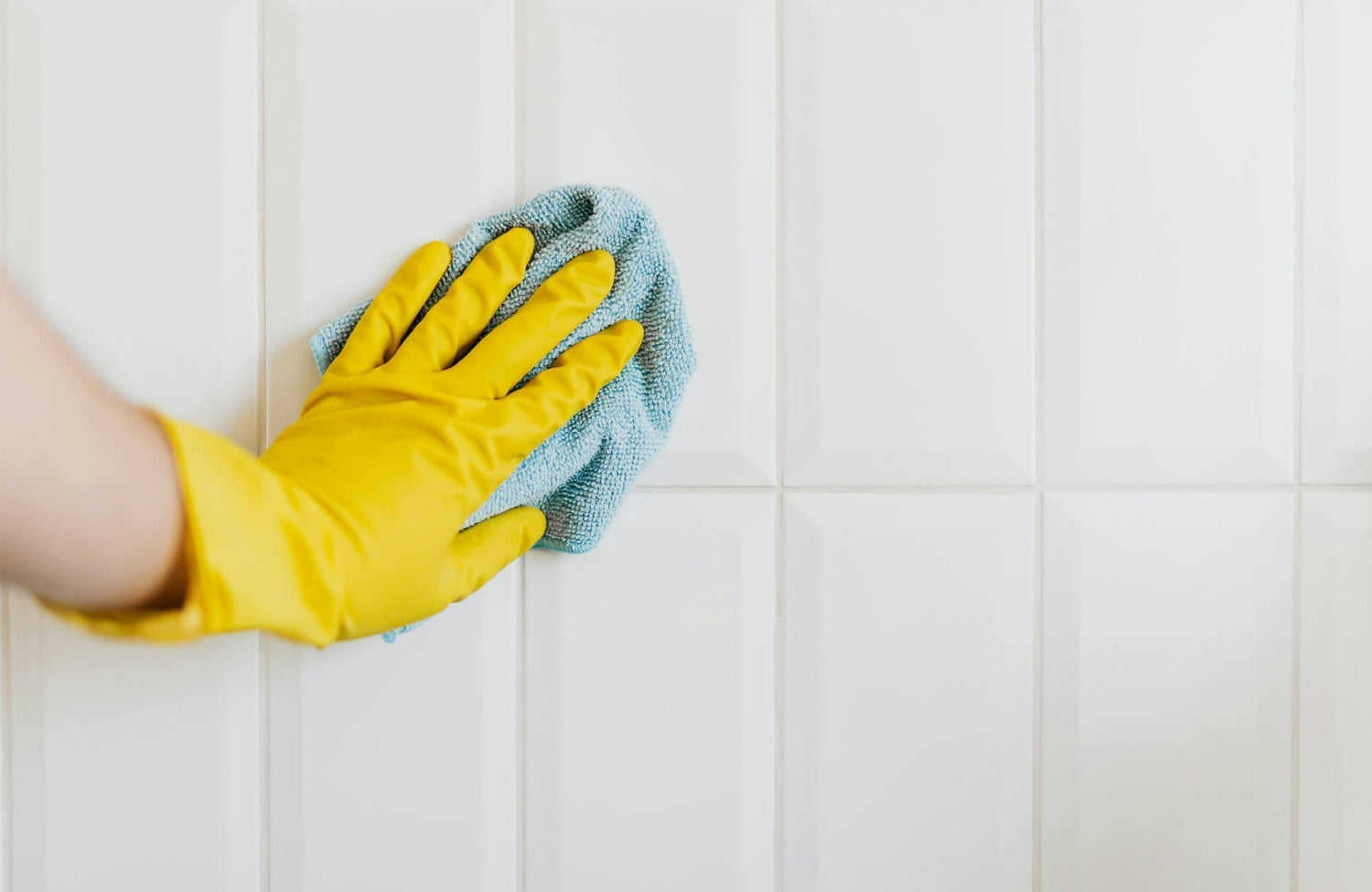 Hand in a yellow glove cleaning glossy white subway beveled tiles with a blue microfiber cloth, highlighting easy maintenance and shine.