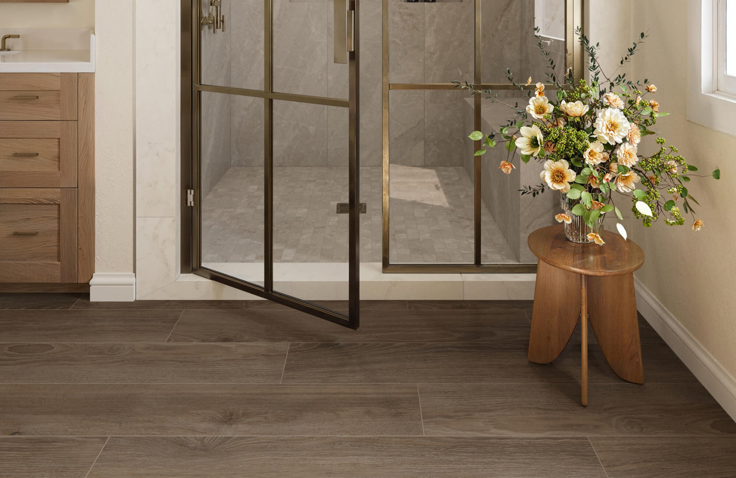 Inviting bathroom with wood-look matte tiles in a rich brown tone, a glass shower with brass framing, and a floral arrangement on a small wooden table, enhancing the natural warmth of the space.