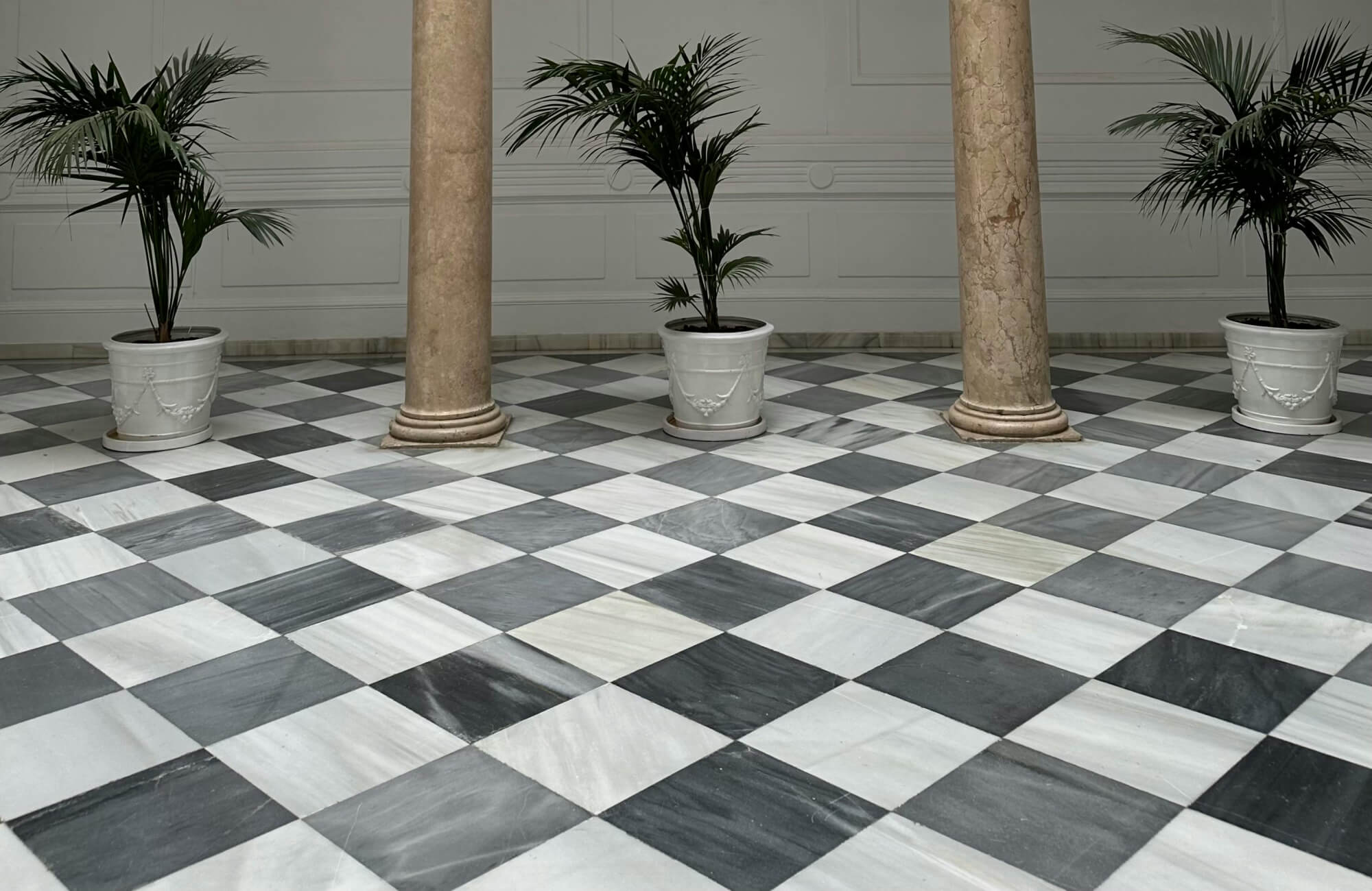 Grand foyer with gray and white checkered floor tiles in a diagonal layout, accented by tall stone columns and lush potted plants for a classic and elegant ambiance