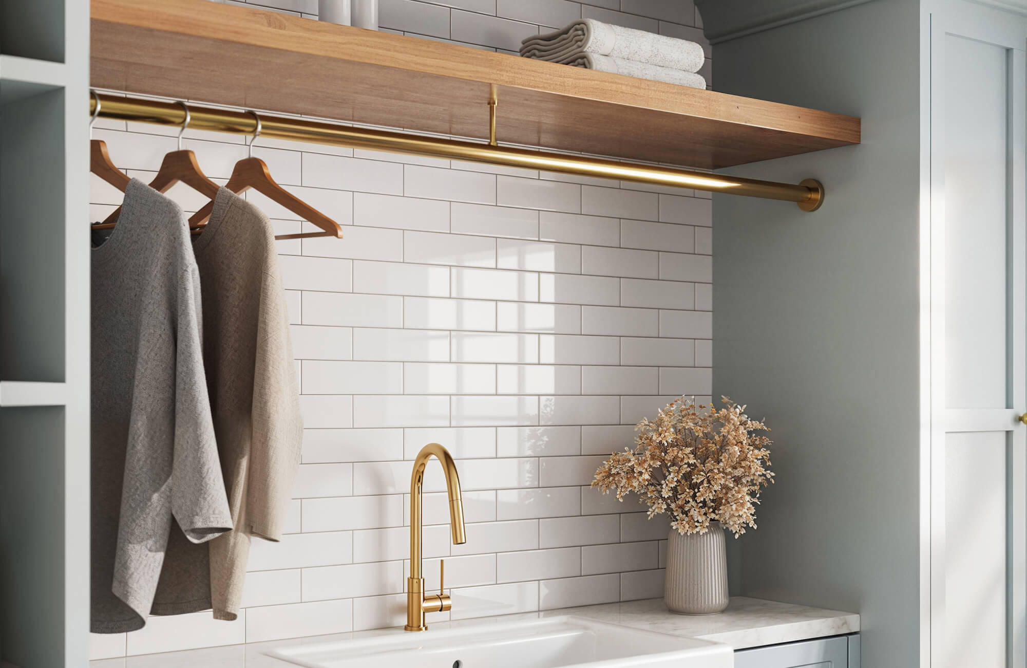 Stylish laundry room featuring glossy white subway tiles, a gold faucet, and elegant decor with hanging clothes and a vase of dried flowers.