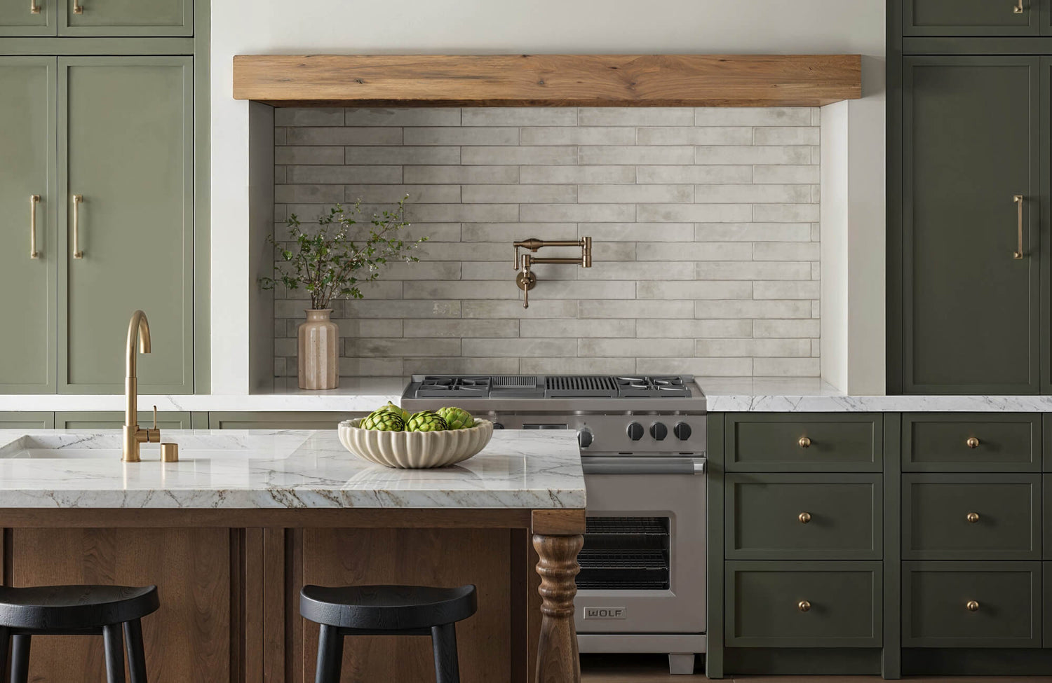 Rustic kitchen featuring gray subway tile backsplash, sage green cabinetry, and a marble island with black stools, accented by brass fixtures and a bowl of artichokes