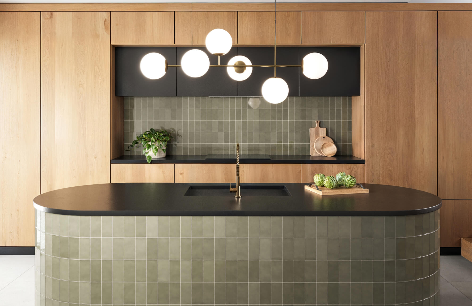 Modern kitchen with a sleek olive green tiled backsplash, natural wood cabinetry, and a curved island featuring matching tiles, highlighted by chic globe lighting.