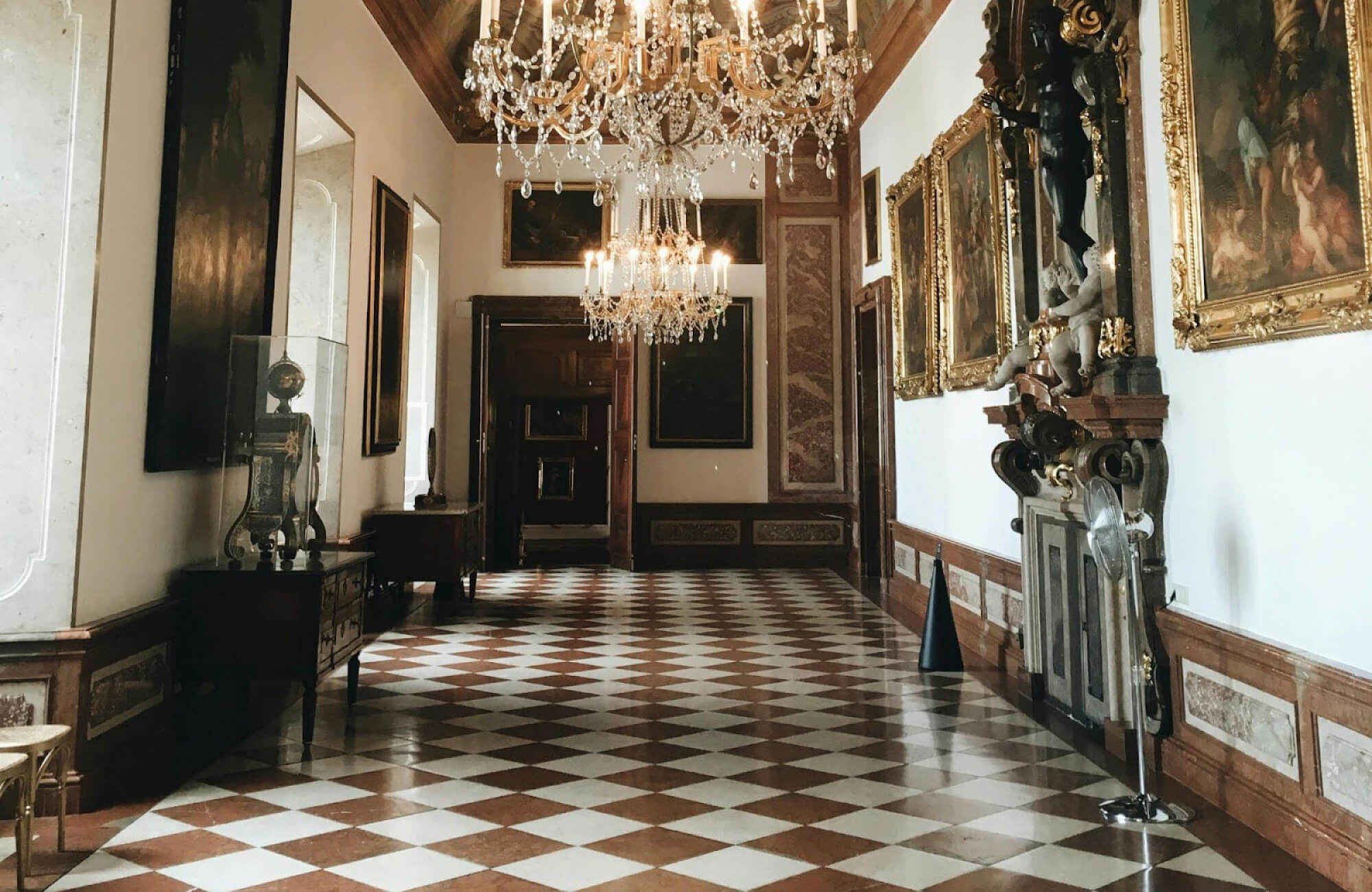 Elegant hallway with polished brown-and-white checkerboard tiles, ornate chandeliers, and gold-framed artwork lining the walls.