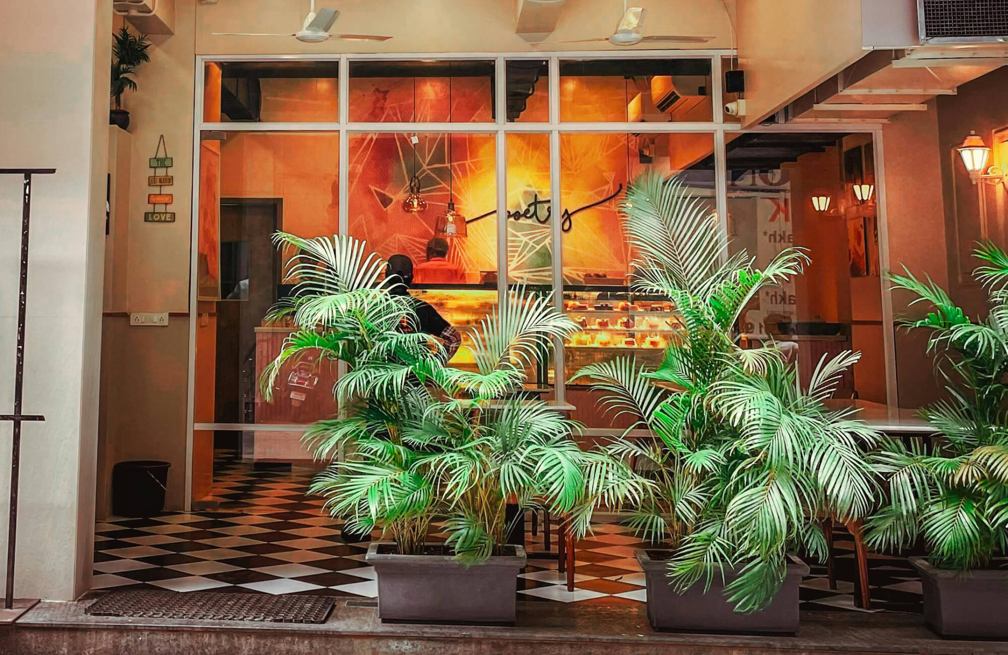 Vibrant café entrance with lush potted plants and black-and-white checkered floor tiles, leading into a warmly lit, modern interior.