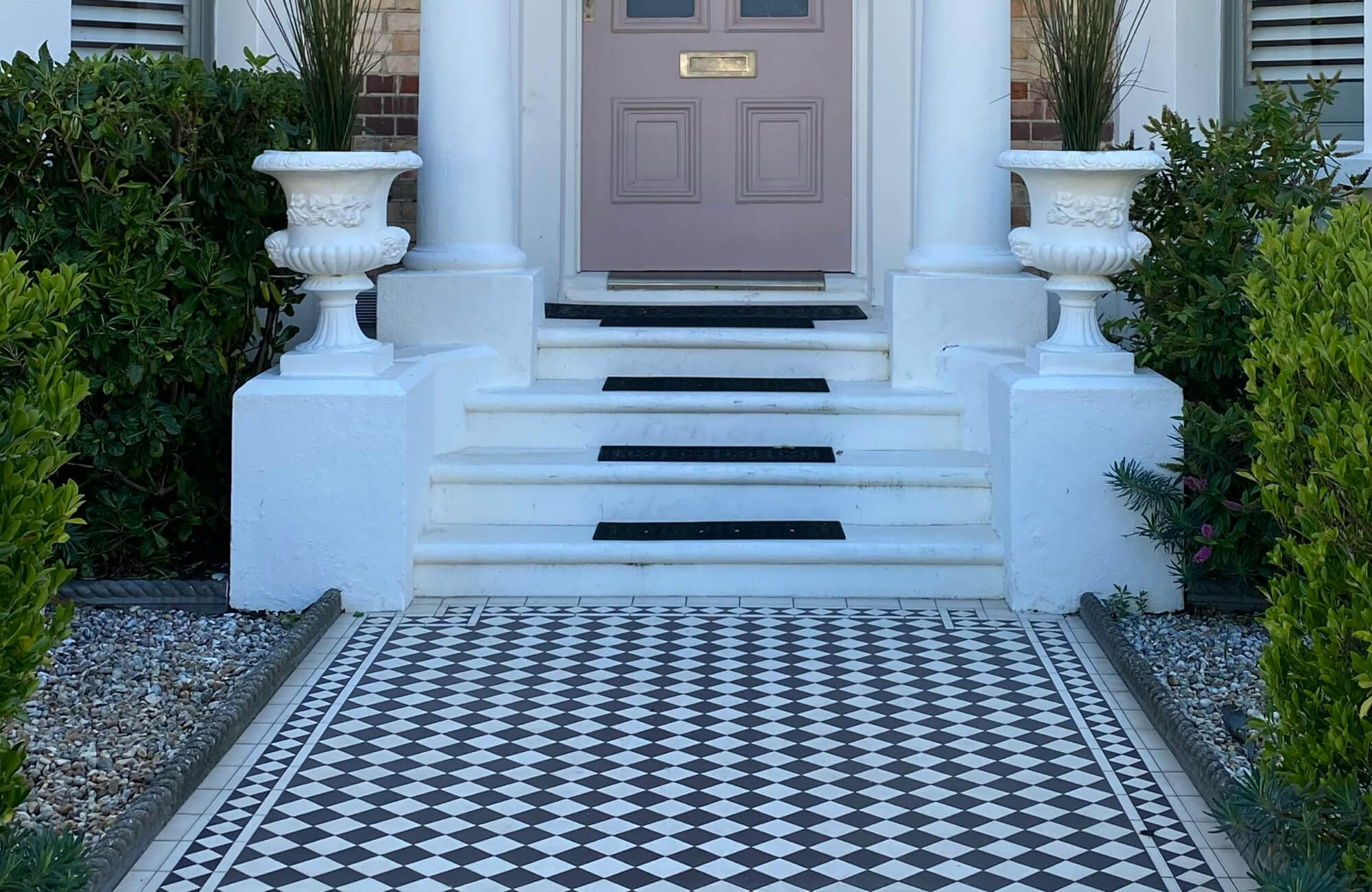 Ornate black-and-white checkerboard tiles line the front pathway of an elegant home, bordered by manicured greenery and classic white planters.