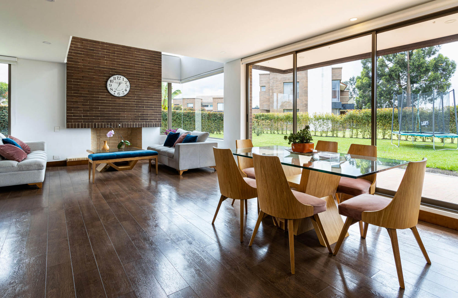 A stylish open living and dining space with dark wood flooring, a brick fireplace, glass dining table, and expansive garden views.