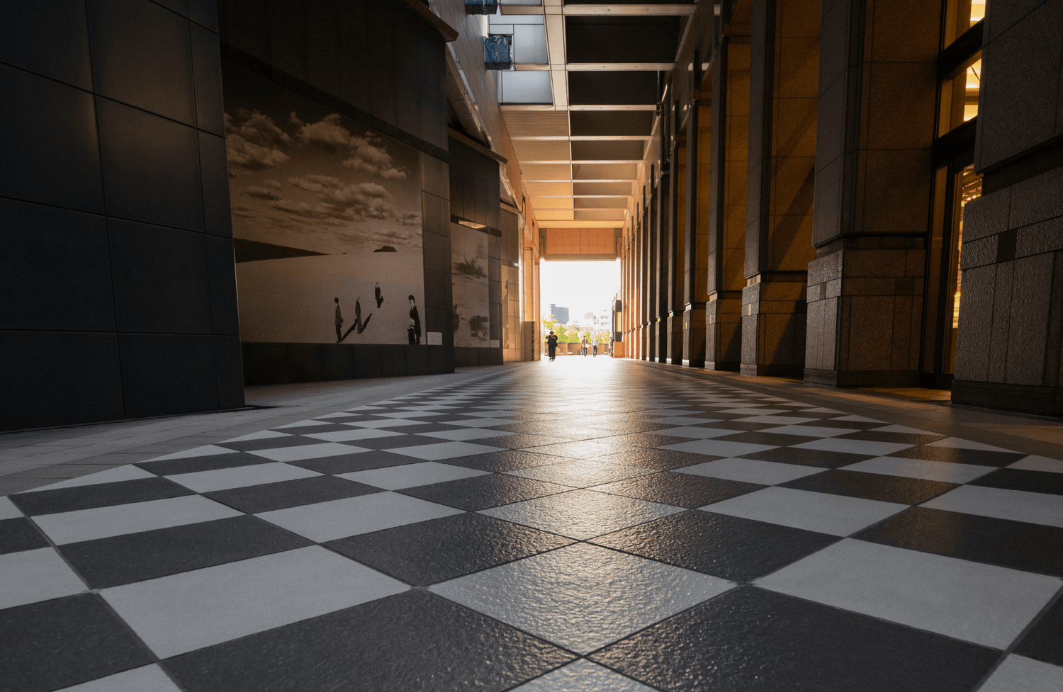 Long hallway with a black and white checkerboard floor, lined with columns and illuminated by warm lighting, leading to an open outdoor space with city views.