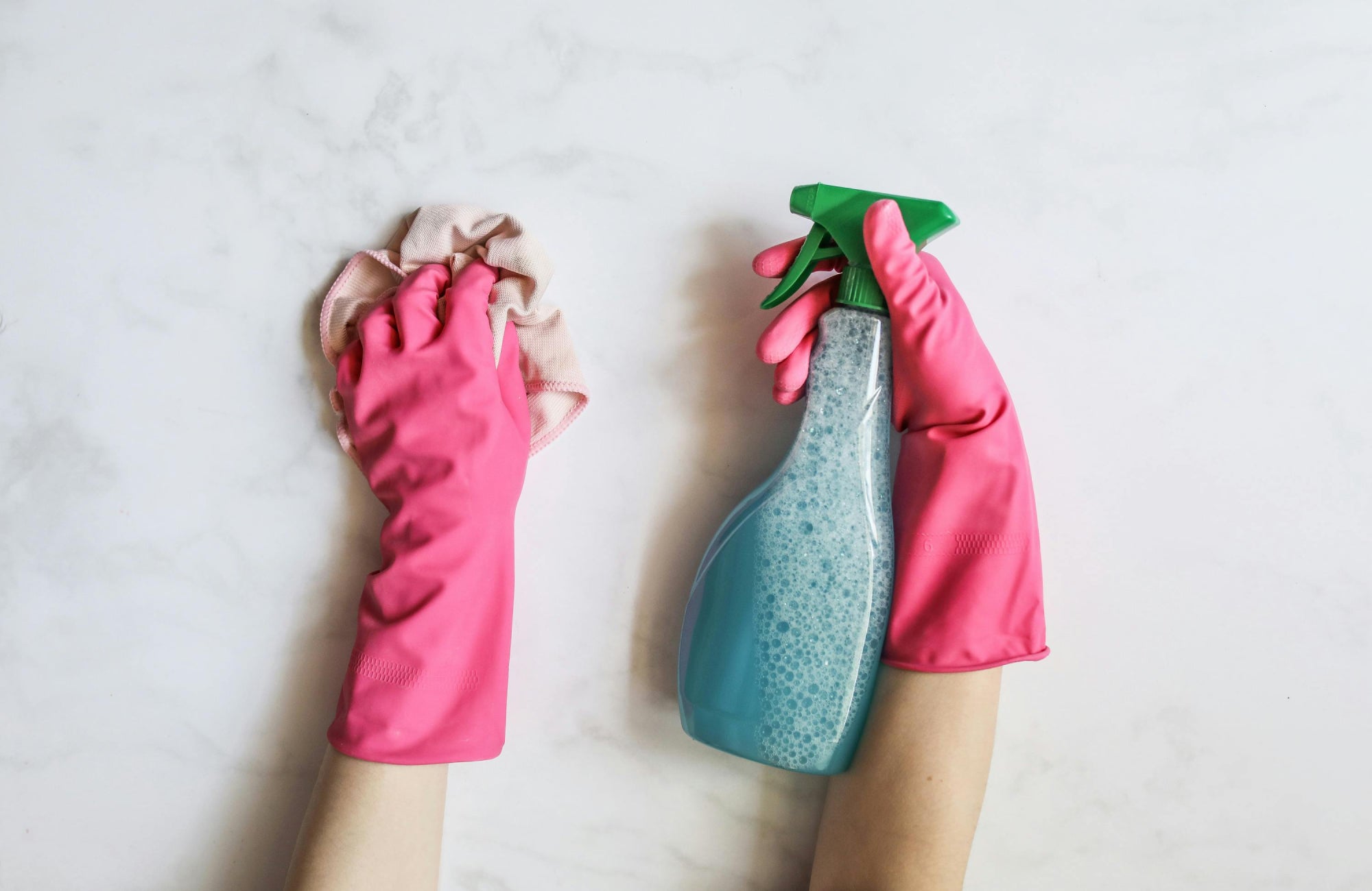 Hands wearing vibrant pink rubber gloves, holding a cloth and a spray bottle with foamy cleaner, poised to clean a white marble surface.