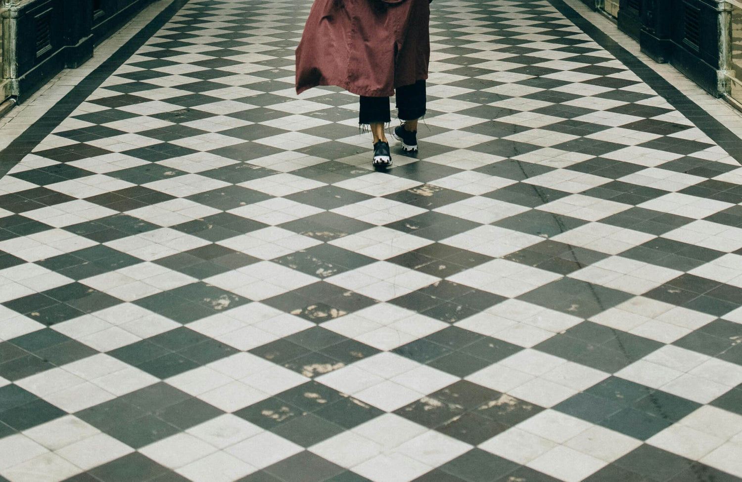 Chipped and scratched black and white checkered floor tiles with a person in a long coat walking, showcasing a classic and timeless design.