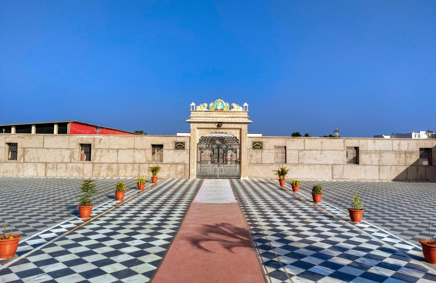 Grand entrance with intricate metal gates framed by beige stone walls, featuring a vast courtyard adorned with classic black-and-white checkerboard tiles and potted plants lining the walkway.