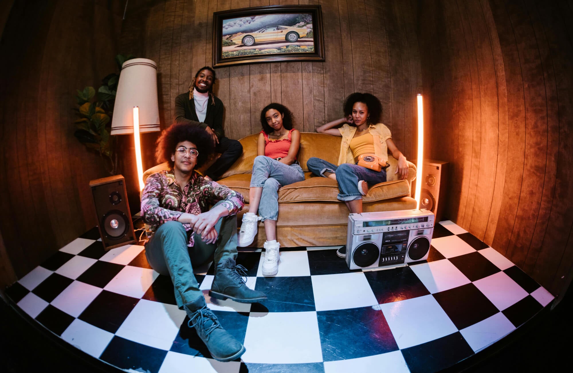 Retro-inspired living room with a checkered black-and-white tile floor, cozy mustard couch, vintage decor, and a group of friends posing with a boombox.