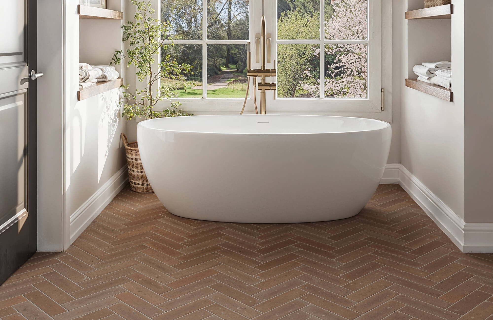 Bright bathroom with a freestanding tub, herringbone-patterned brown subway tile flooring, and large windows overlooking a lush garden.