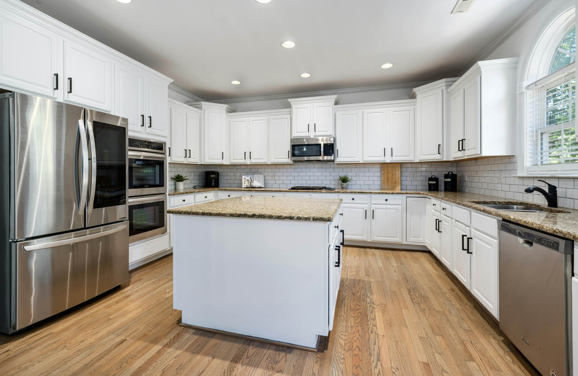 A spacious kitchen with white cabinetry, stainless steel appliances, and warm hardwood flooring, centered by a granite-topped island.