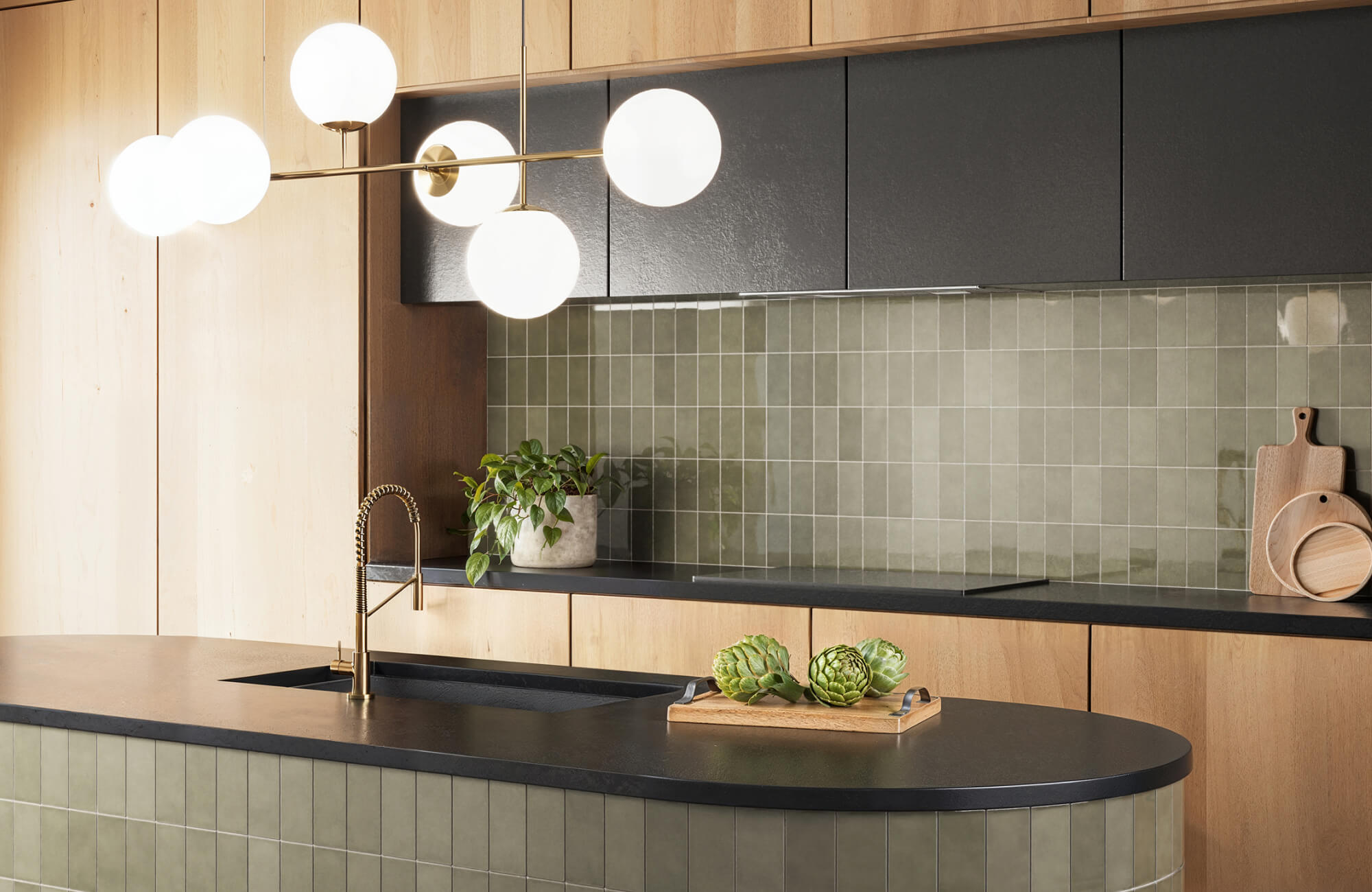 Contemporary kitchen with matte black countertops, sage green subway tile backsplash, and natural wood cabinetry, accented by a modern globe light fixture.