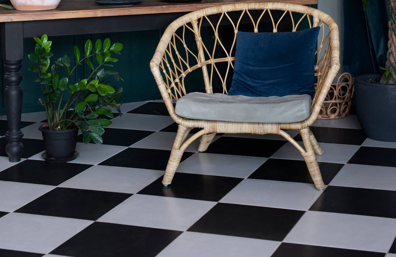 Cozy seating area with black-and-white checkered porcelain floor tiles, featuring a wicker chair, wooden table, and vibrant green potted plants for a natural touch.