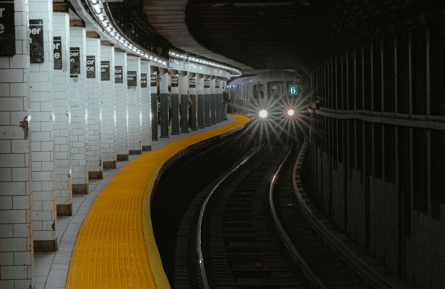 A New York subway train approaches a station with white subway tiles lining the curved walls and a bright yellow tactile paving along the platform edge