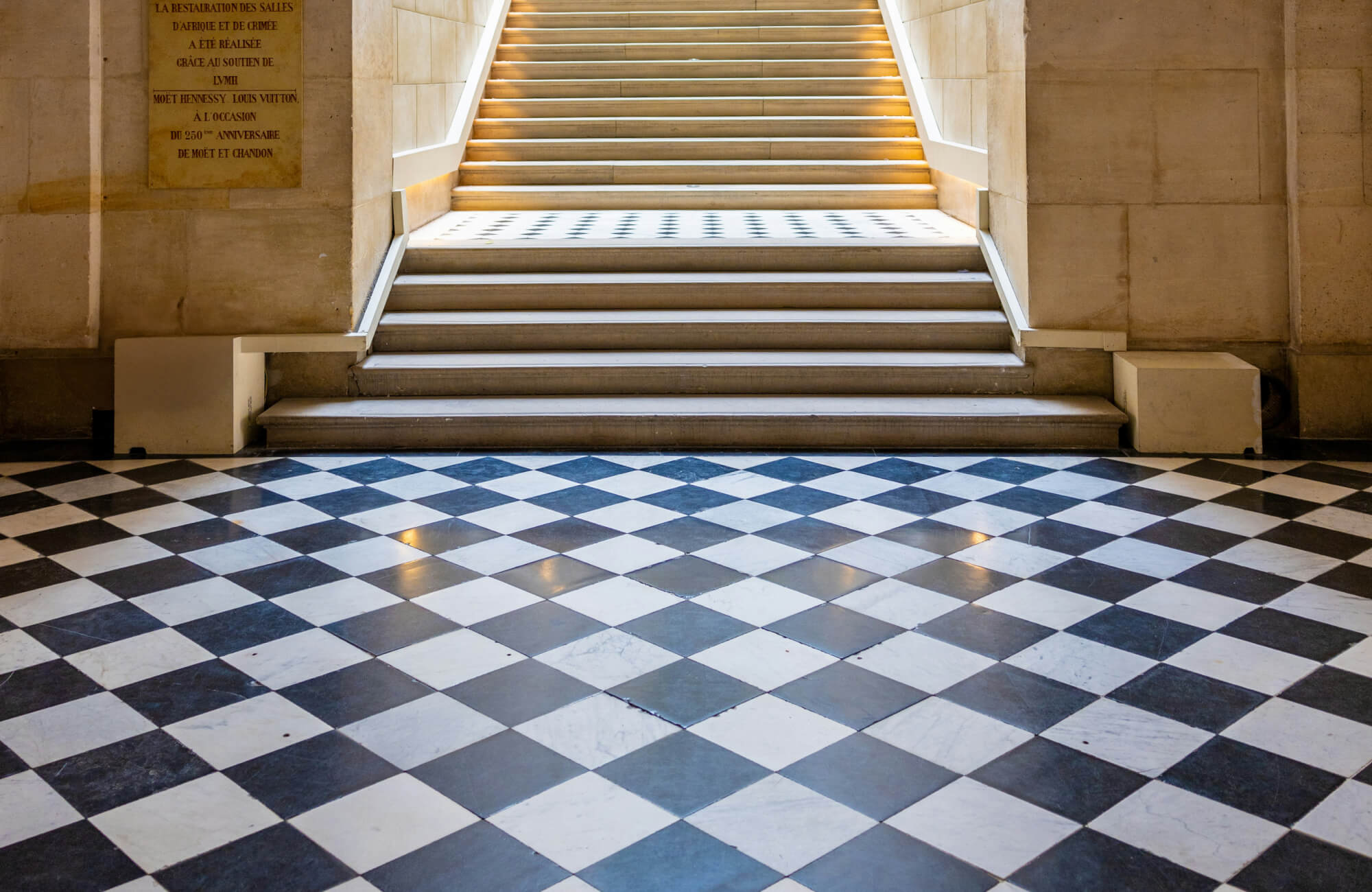 Grand staircase leading to a hallway with a black and white checkerboard tile floor, surrounded by beige stone walls