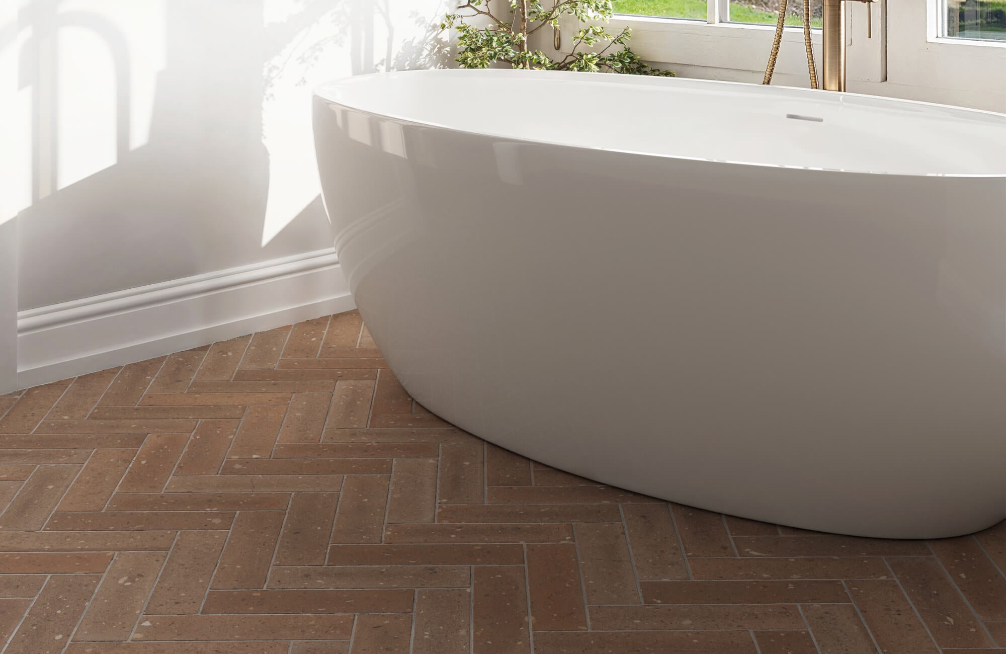 Luxurious bathroom featuring a white freestanding tub on a floor with brown subway tiles arranged in a classic herringbone pattern, basking in soft natural light from the windows.