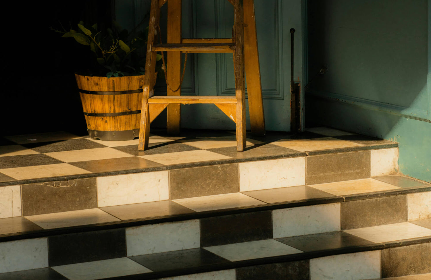 A rustic staircase with black and white chequered tiles, a wooden ladder, and a wooden bucket plant under warm sunlight