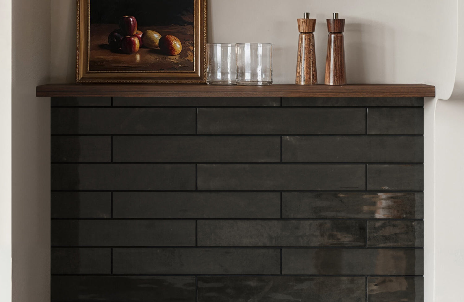 A sleek, modern fireplace adorned with matte black subway tiles, topped with a wooden mantle showcasing a framed still-life painting and rustic decor.