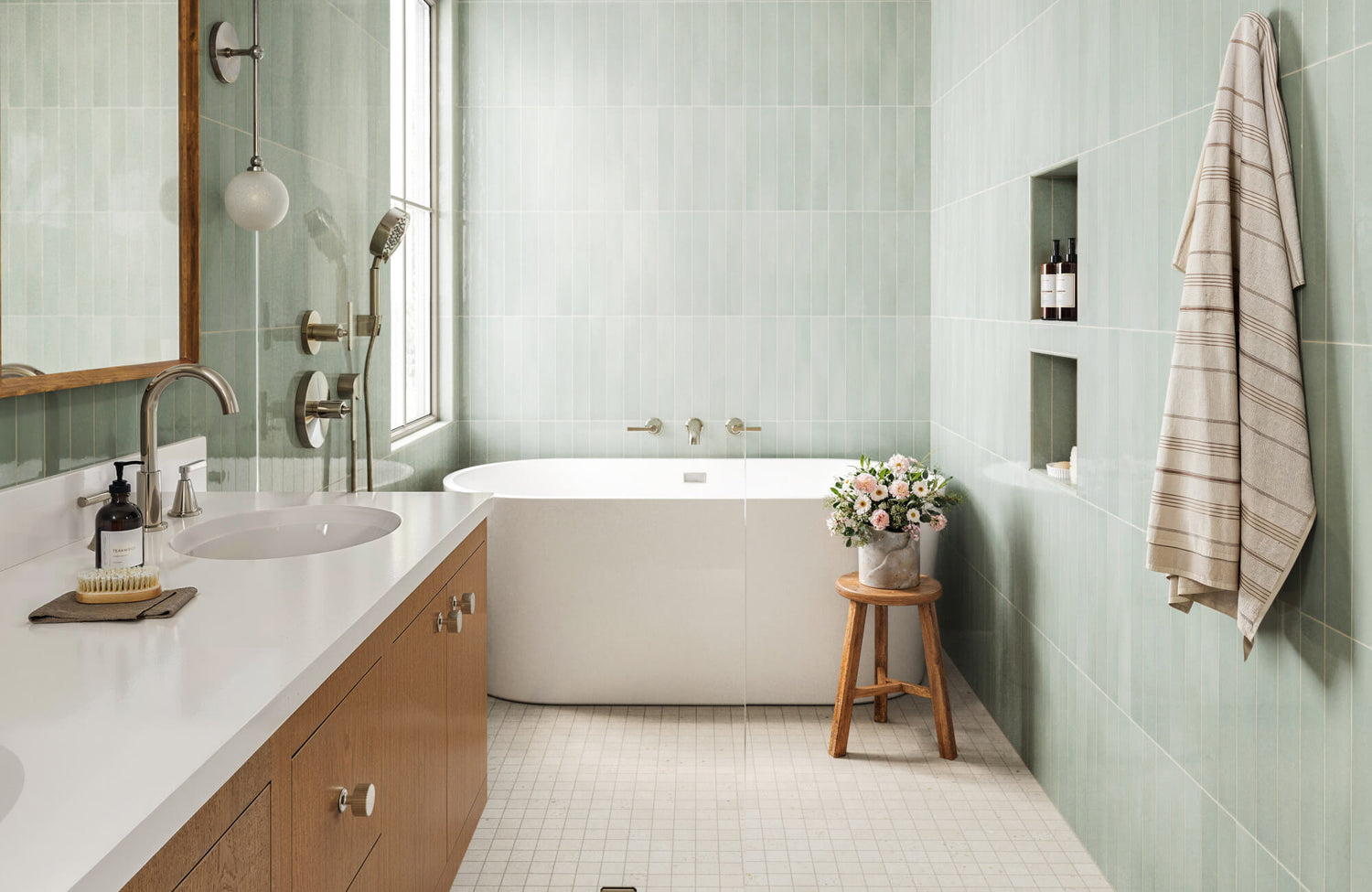 Serene bathroom with soft green subway tiles, a sleek freestanding tub, and a warm wood vanity accented by fresh flowers.