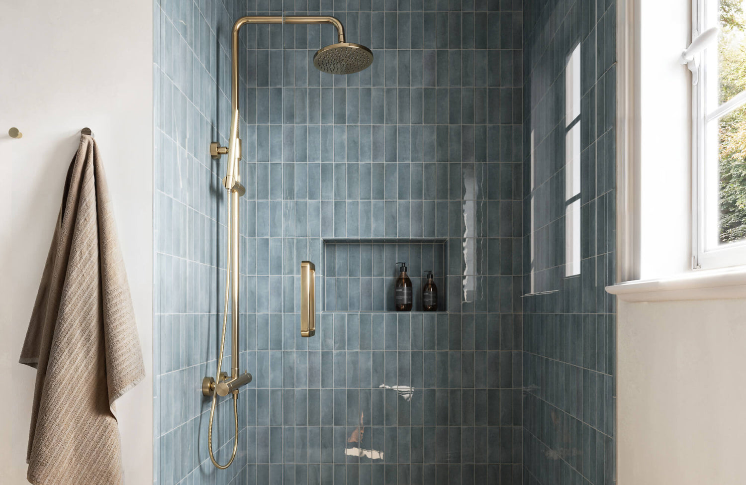 Elegant teal subway tiles with a glossy finish line the shower walls, complemented by a brass rain showerhead and built-in shelf for a refined, modern look.