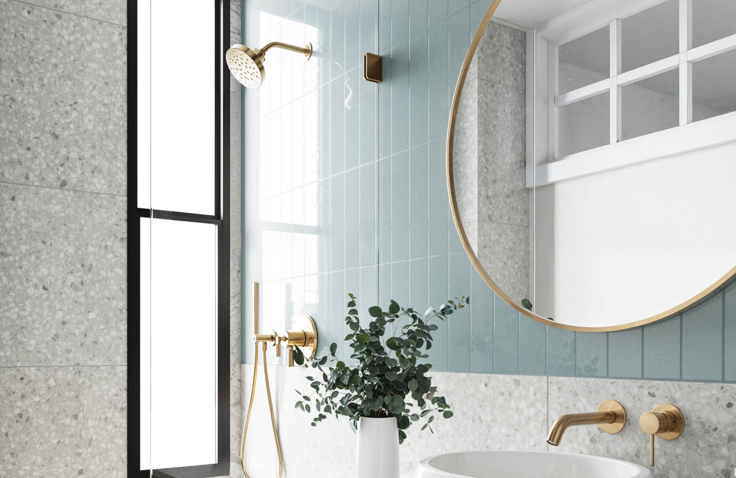 Elegant bathroom with soft blue subway tiles, terrazzo wall, and gold fixtures, featuring a round mirror and minimalist decor.