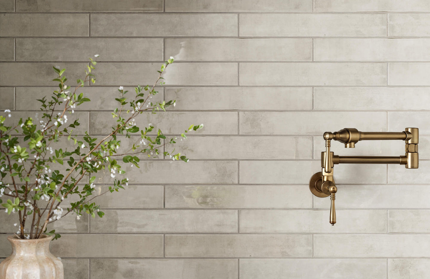 Elegant kitchen wall featuring elongated subway tiles in muted beige with a vintage brass pot filler and a vase with flowering branches.