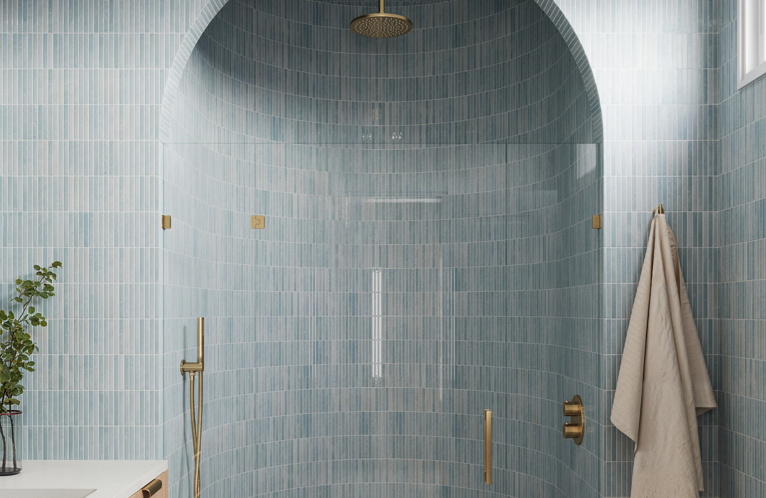 An elegant bathroom with vertical light blue tiles and brass fixtures, featuring a curved walk-in shower and a hanging towel.