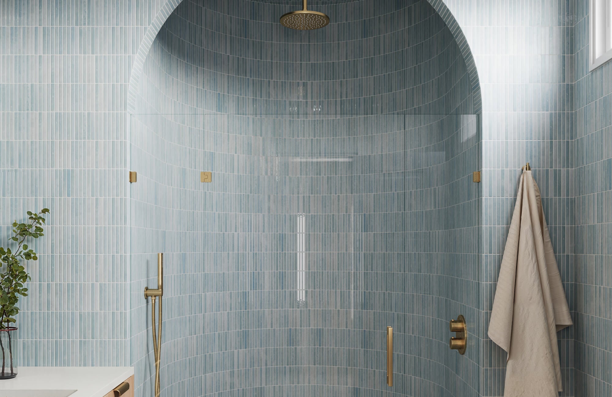 An elegant bathroom with vertical light blue tiles and brass fixtures, featuring a curved walk-in shower and a hanging towel.