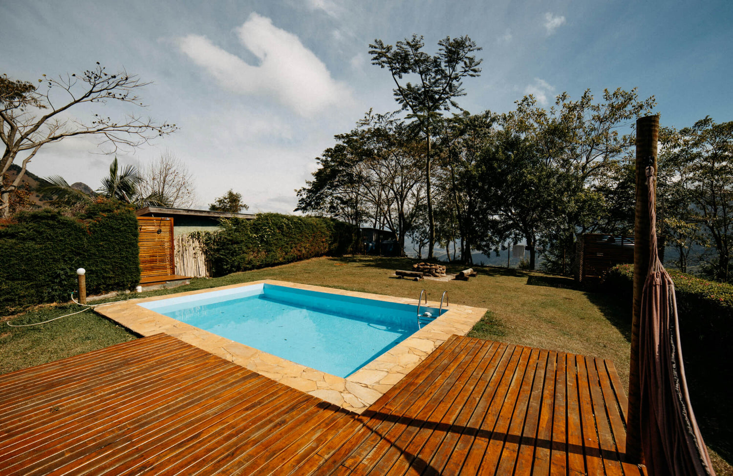 Charming outdoor space featuring a vibrant blue pool surrounded by natural stone tiles and warm wooden decking, set amidst lush greenery.