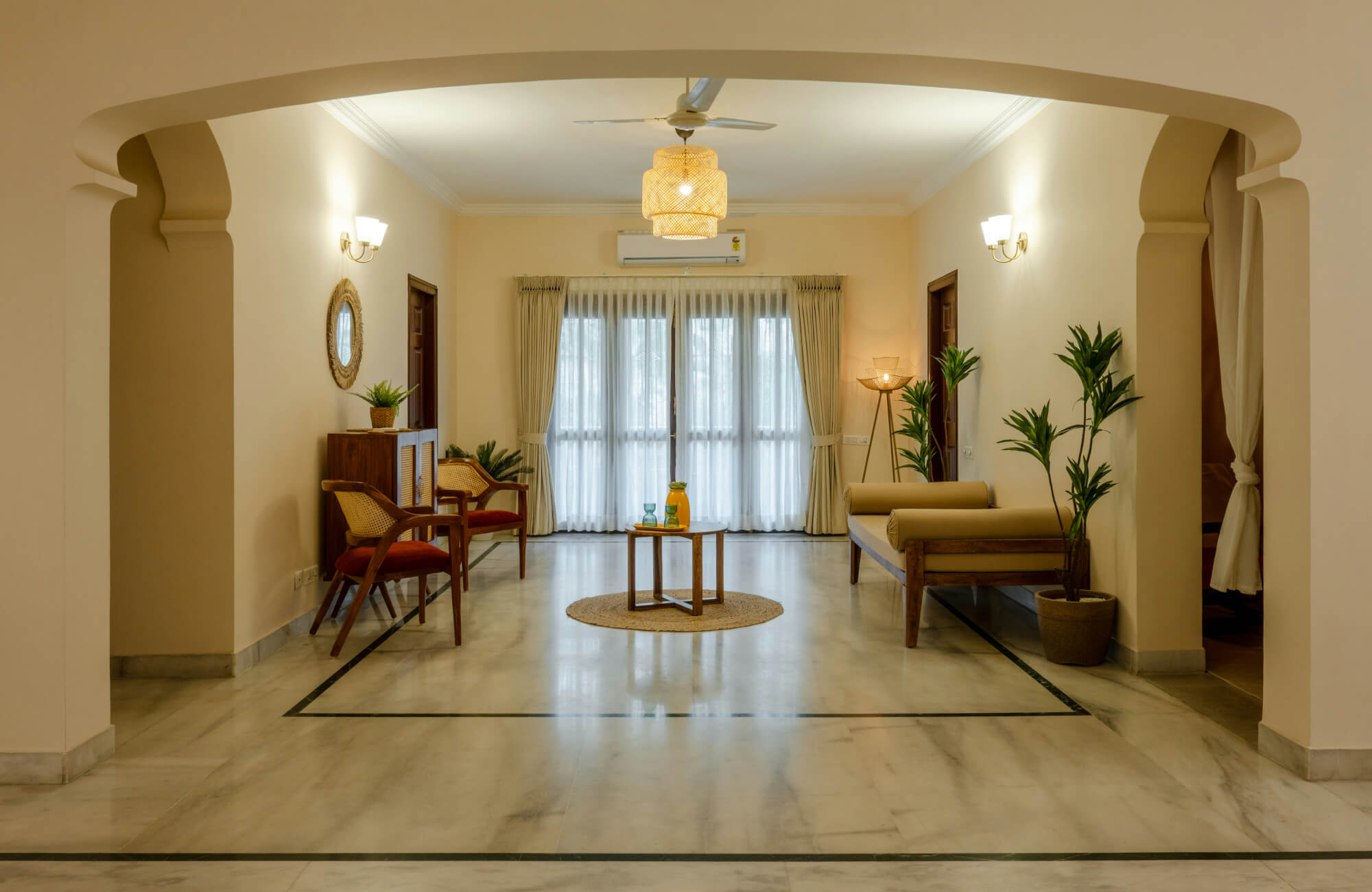 Spacious living room with polished marble floors, light cream walls, and wooden furniture. Large windows with sheer curtains provide natural light. 