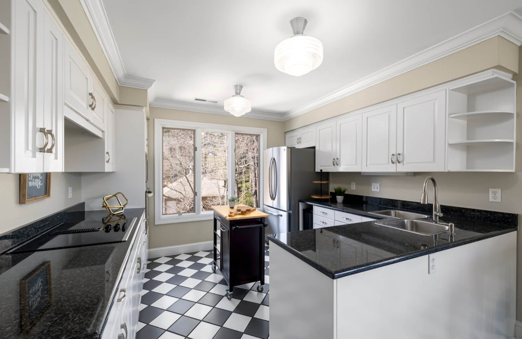 Bright kitchen with black and white checkered floor, white cabinets, and sleek black countertops, featuring a central island and large window.