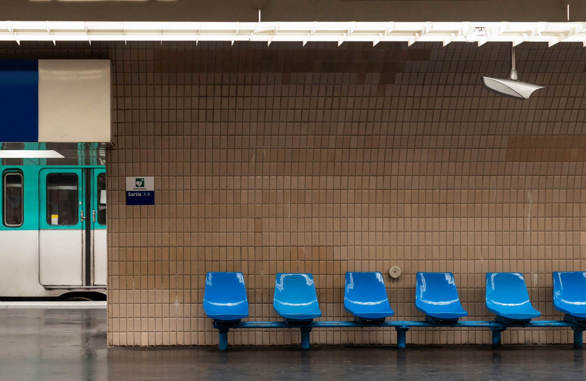 A subway platform with beige tile walls, blue plastic seats, and a teal metro train in the background.