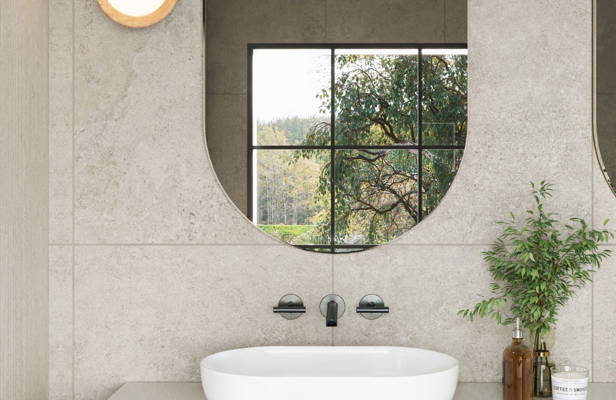 Modern bathroom featuring large format stone look bathroom tiles with a textured finish, a sleek round mirror, and matte black fixtures, complemented by natural greenery.