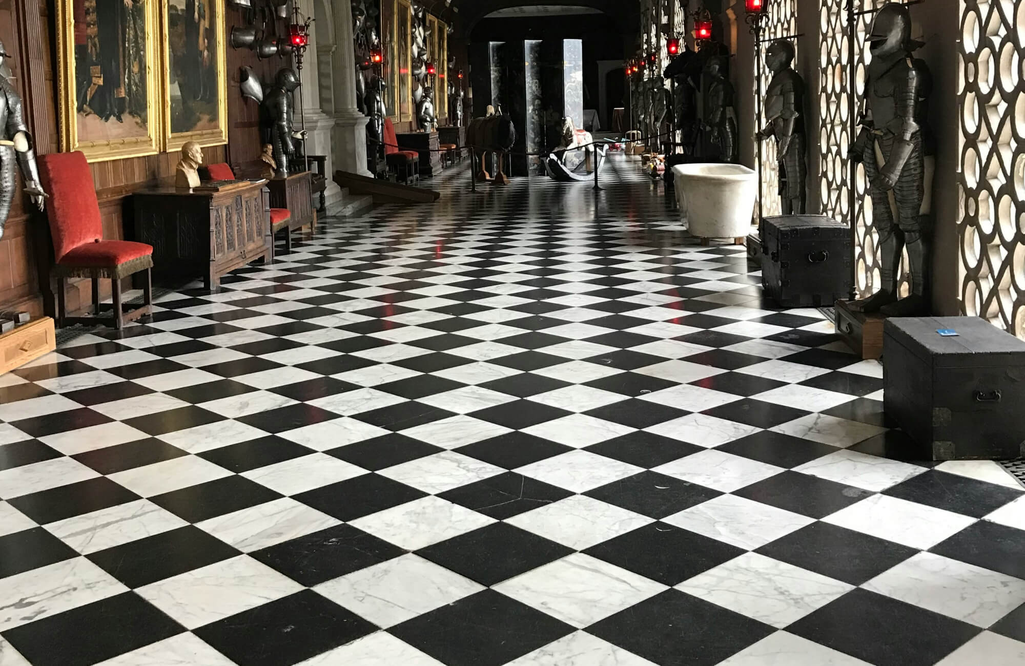 Grand corridor with black and white marble look checkered floor tiles, flanked by medieval armor and red velvet chairs, exuding historical elegance