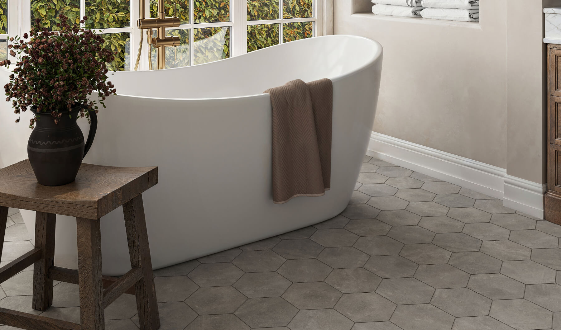 Elegant hexagonal dark grey tiles pave the floor, complementing a freestanding white tub accented by a rustic wooden bench and lush potted blooms