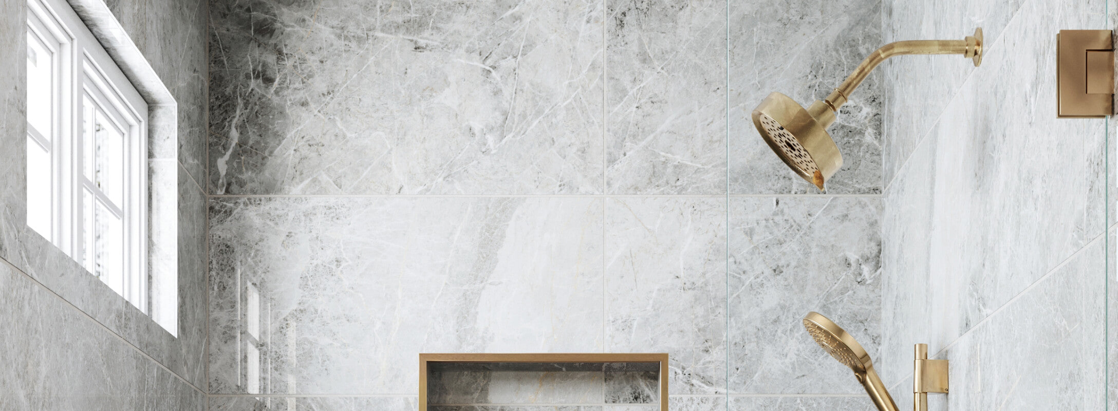 Luxurious shower with grey marble look tiles and dual brass showerheads, complemented by a built-in shelf and a window for natural light