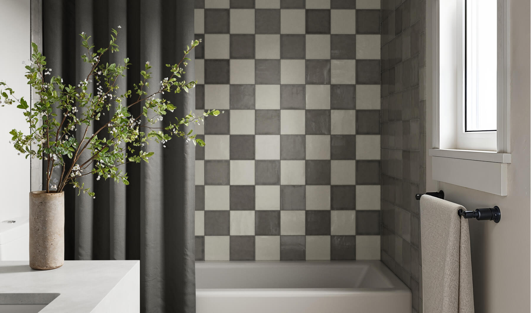 Modern bathroom featuring checkerboard wall tiles in shades of gray and beige, a sleek white bathtub, and a decorative vase with greenery