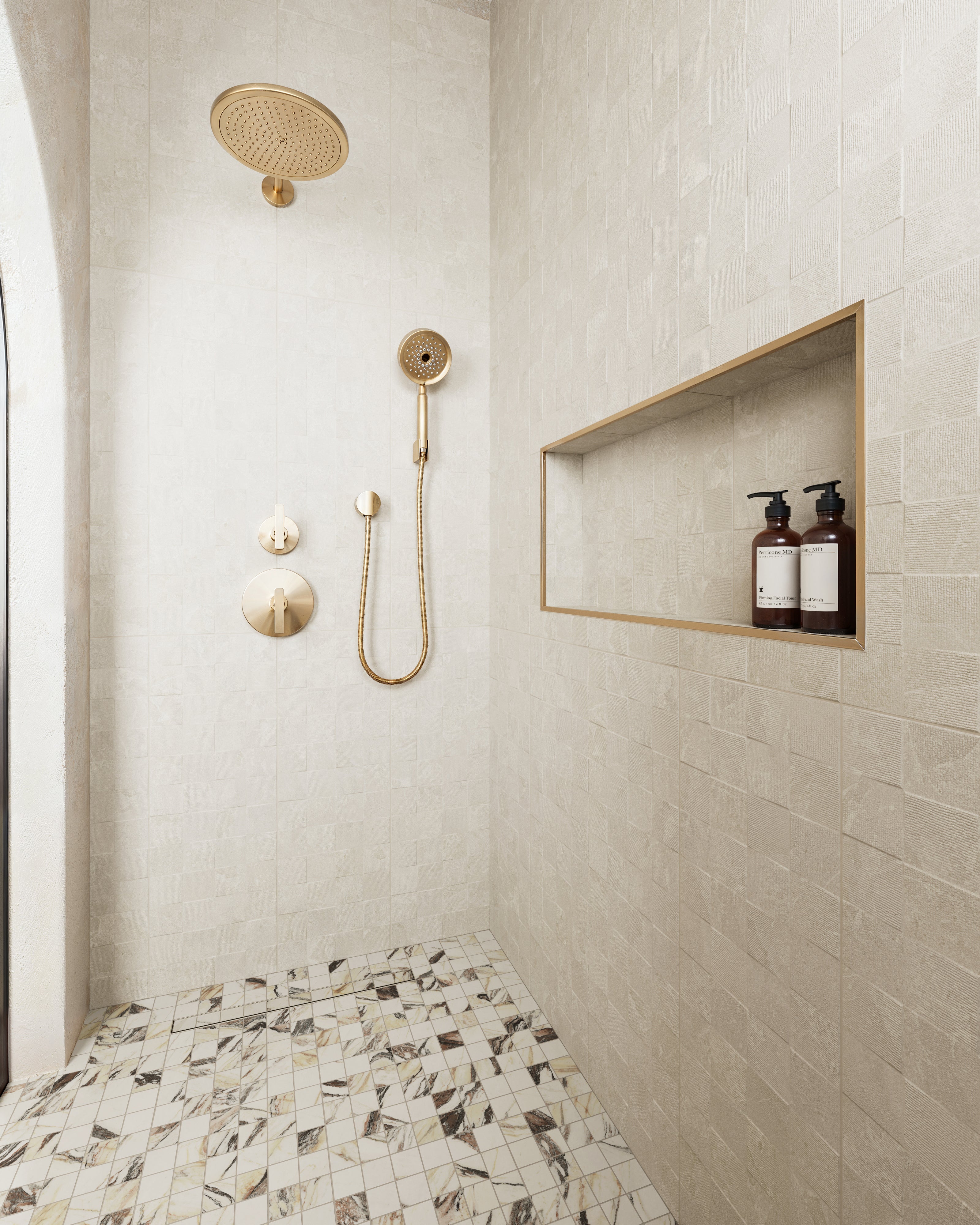 Elegant shower featuring beige textured tiles, marble-look mosaic flooring, gold shower fixtures, and a recessed shelf for added functionality.