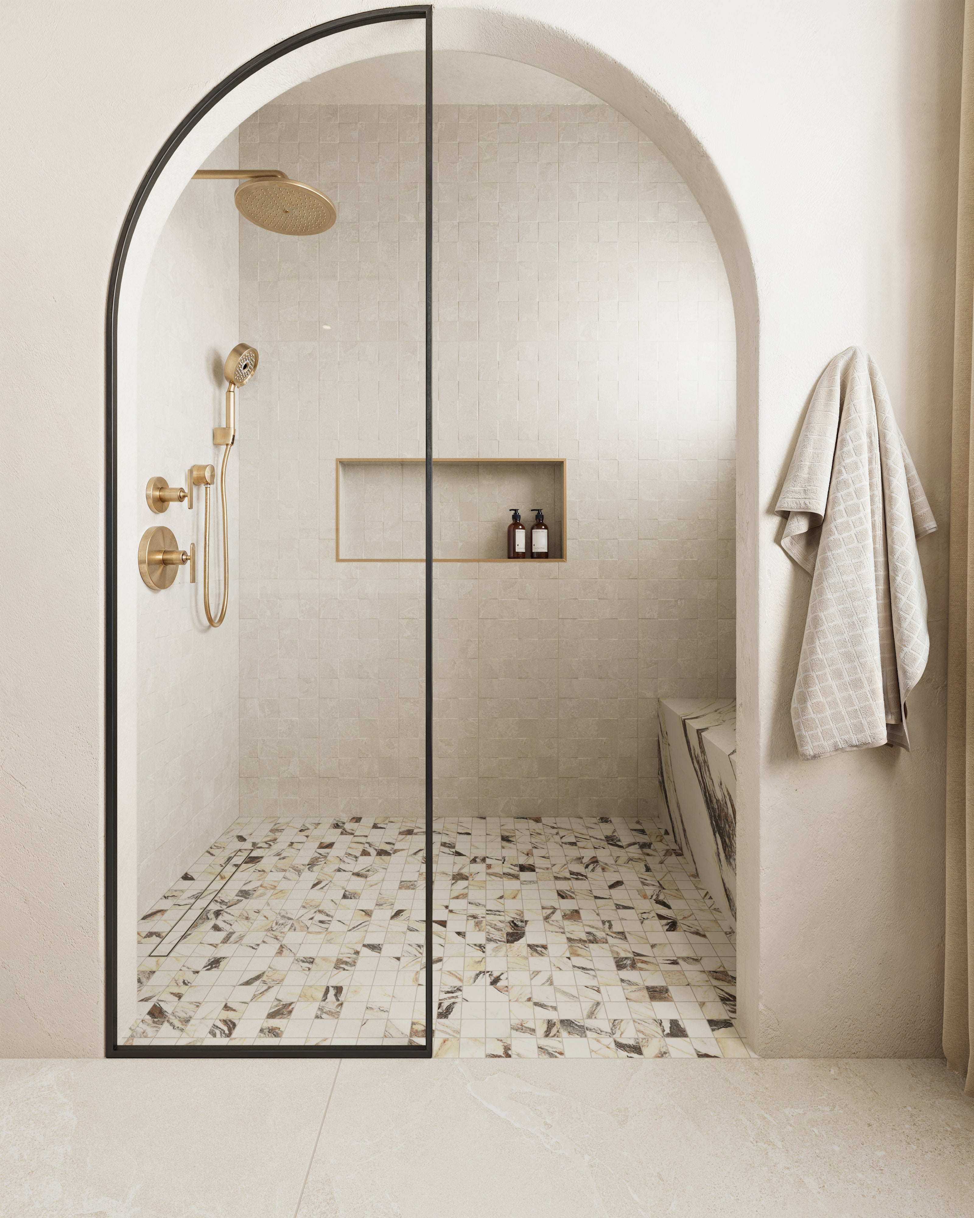 Modern shower with marble-look mosaic flooring, beige decorative wall tiles, arched glass door, gold fixtures, and built-in shelf for toiletries.