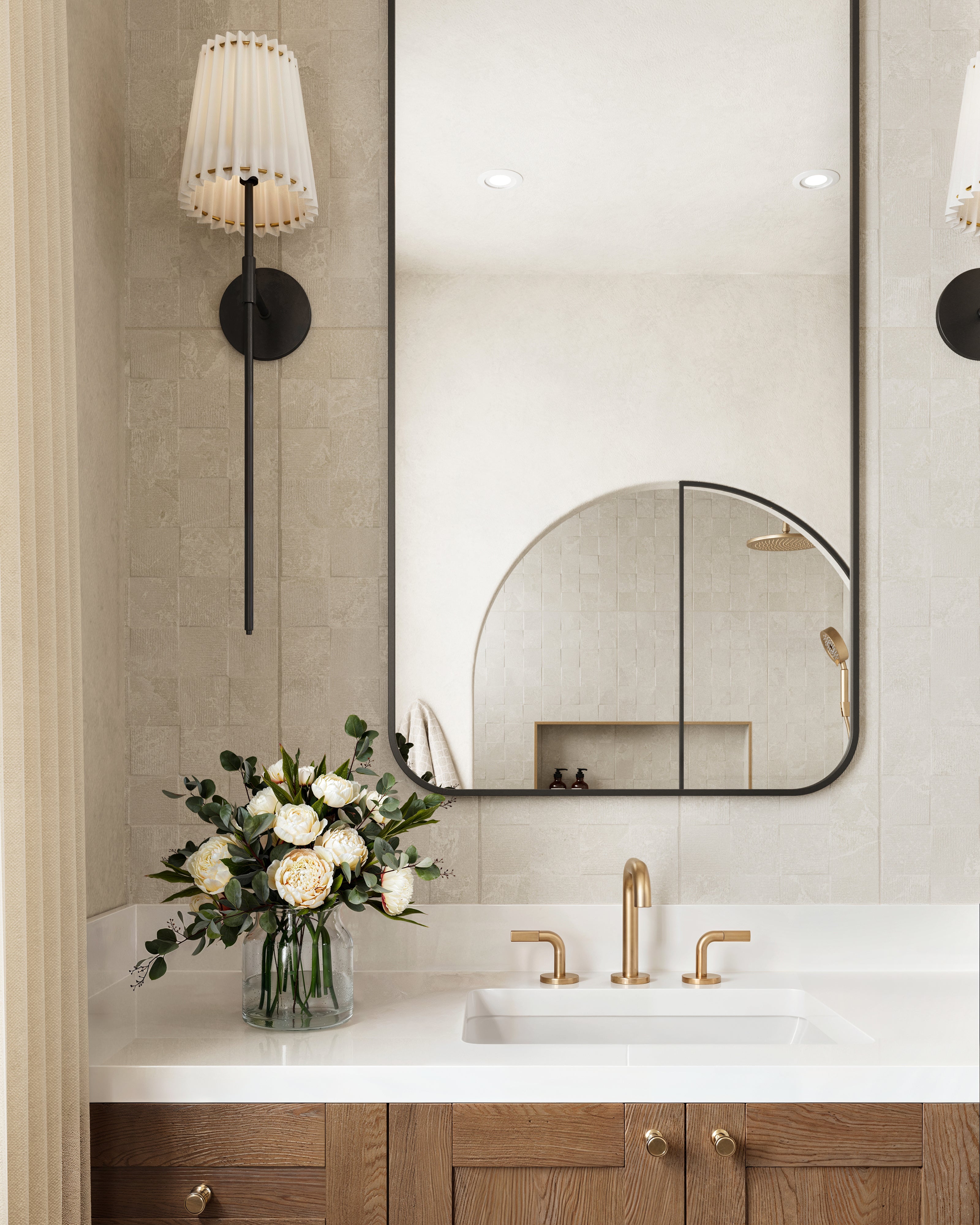 Elegant bathroom vanity with a white countertop, gold faucet, floral arrangement, and beige decorative tiles, reflecting an arched shower in the mirror.