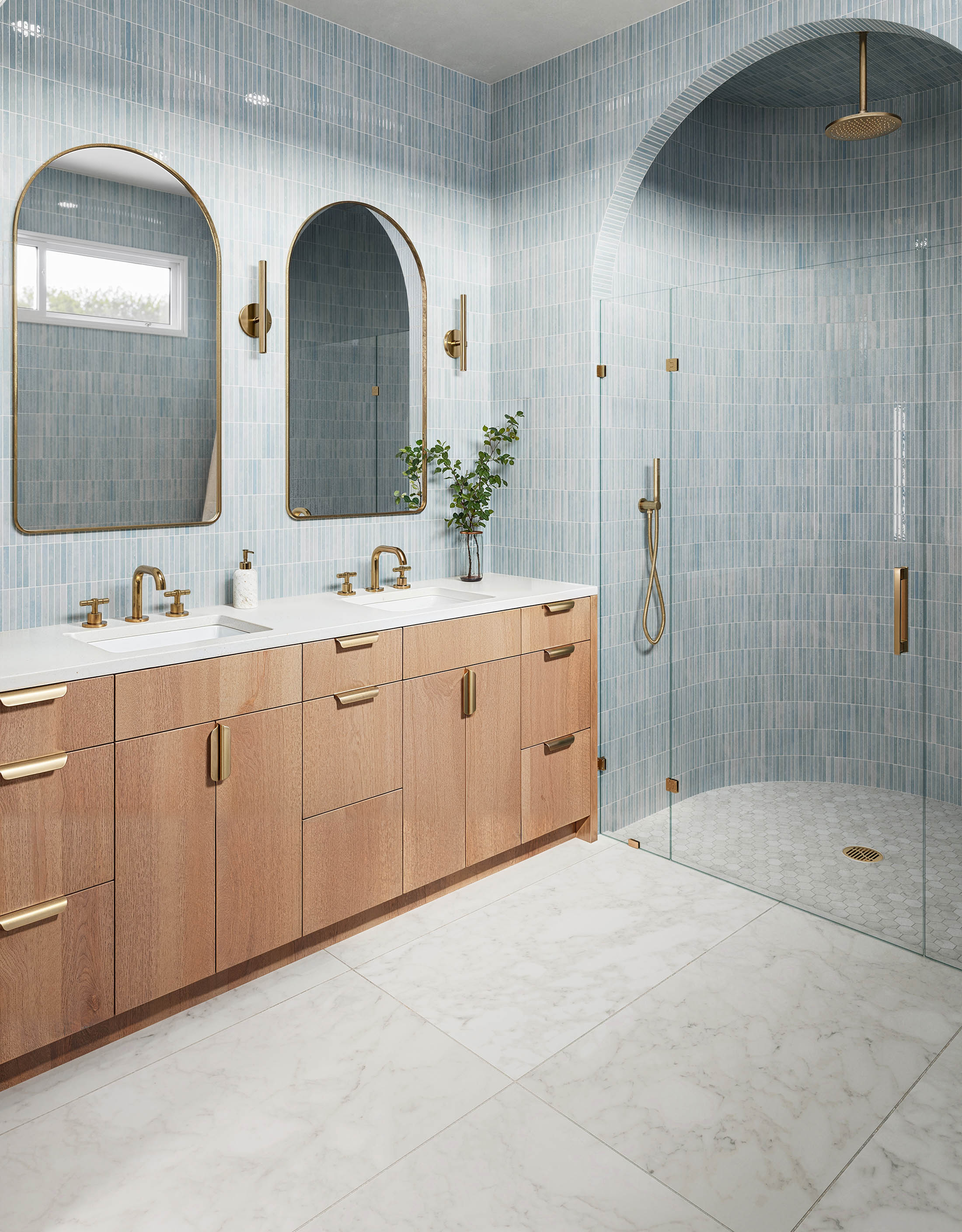 Bright bathroom with glossy blue subway tiles, a double wood vanity with gold hardware, white marble look floor tiles, and a matching arched glass shower.