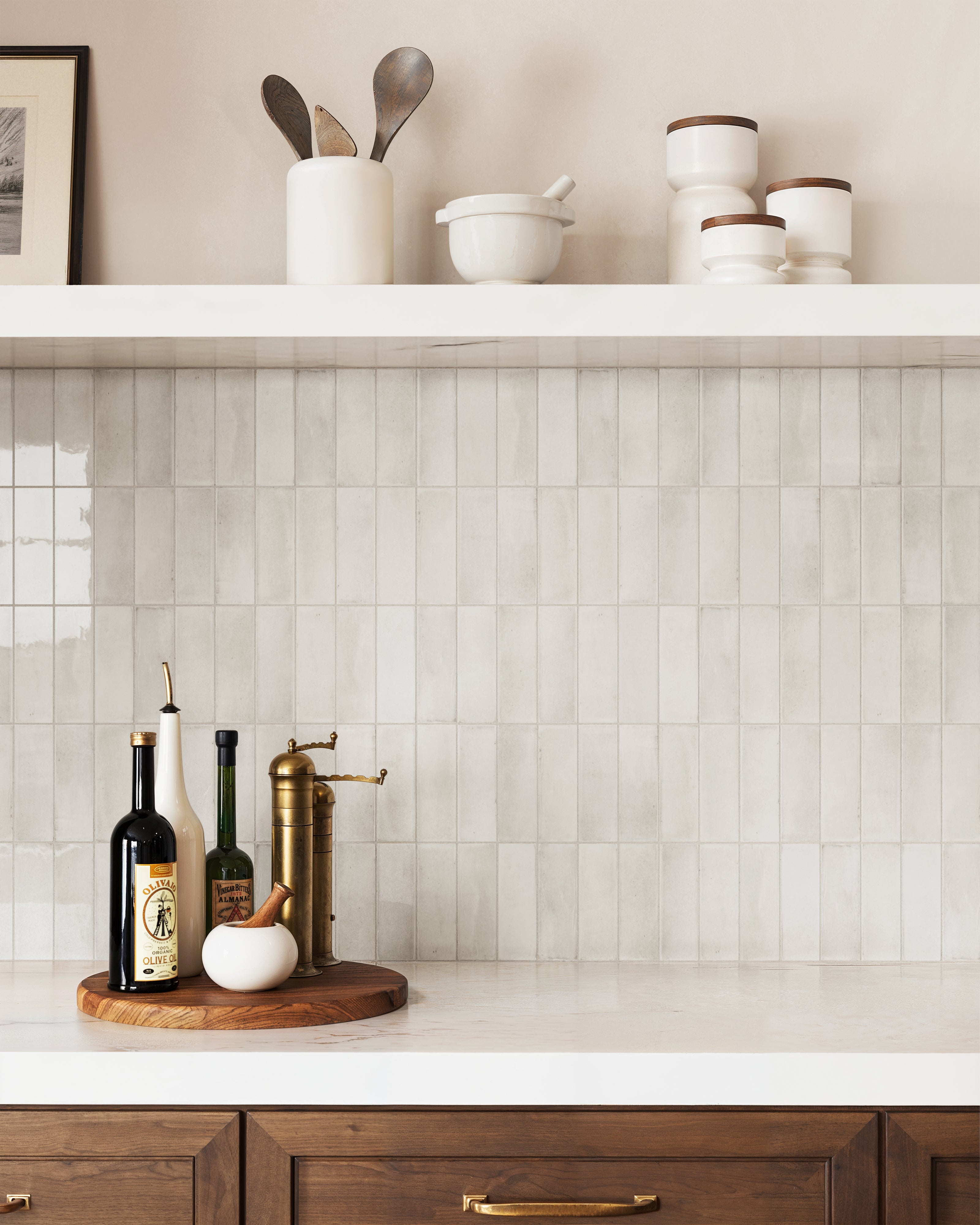 The kitchen backsplash features glossy taupe tiles arranged vertically, adding a subtle texture and warmth that complements the wooden cabinetry and brass accents.